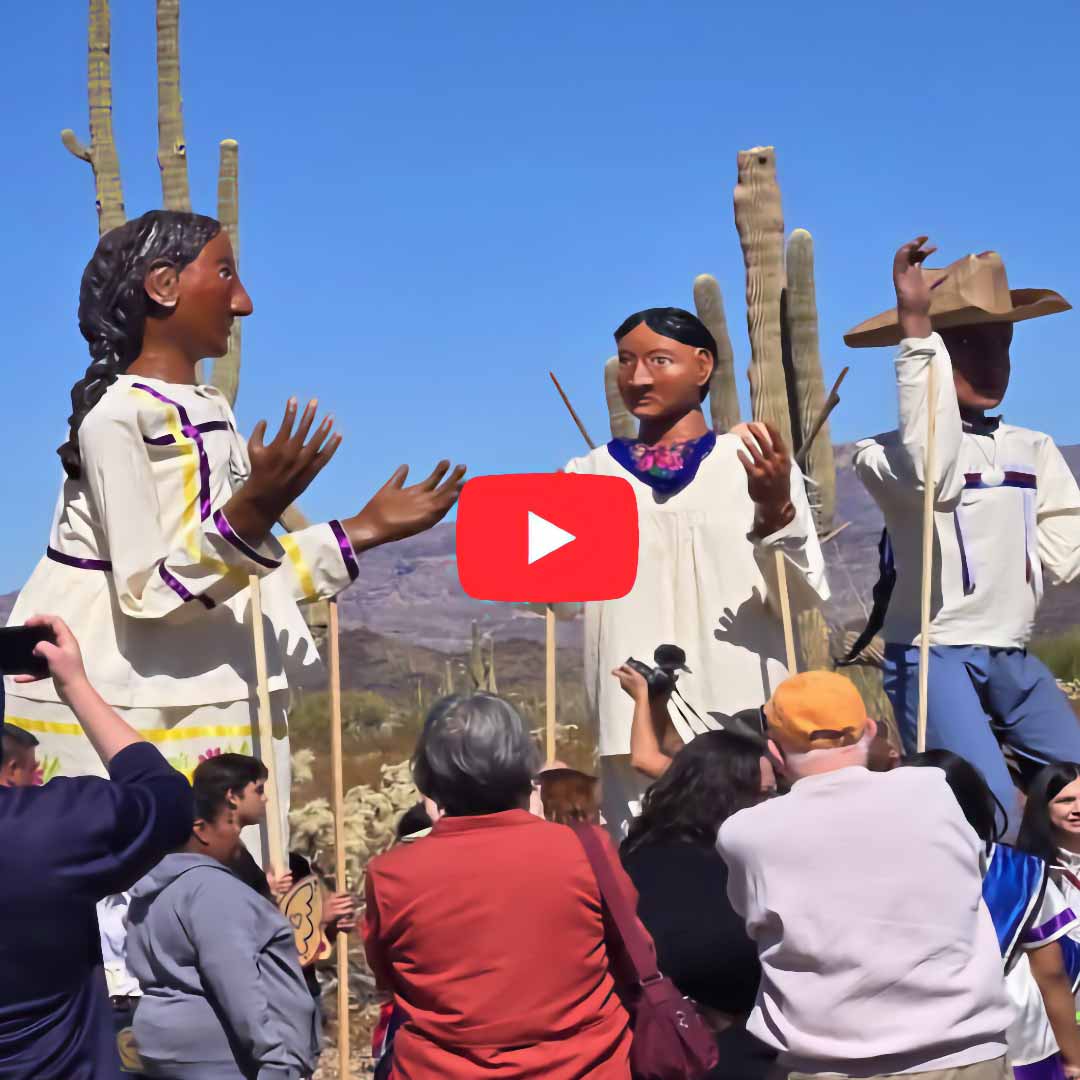 Cropped view of a video still with a red YouTube “play” button in the center. The still shows a group of people gathered outdoors at the Organ Pipe Cactus National Monument in the Sonoran Desert in Arizona. They are collectively supporting three large puppets, each 10 to 15 feet tall, representing two women with brown skin and black hair and man with brown skin and a cowboy hat. Some of the puppeteers hold long sticks attached to the puppets’ wrists in order to make their arms and hands move. Behind the puppets are two tall saguaro cacti, and in the distance are low hills.