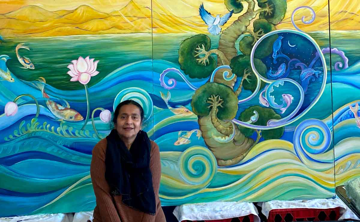 The artist Geeta Dave sits in front of a finished mural that is supported by red plastic crates covered in white cloth. The image is cropped to show only a lower left section of the mural, which is several times larger than shown. In the visible portion, we can see fish, pink lotus flowers, a green sea in the distance, blue and green curving waves in the foreground, a green tree-like structure emerging from the water with branching growths that resemble coral, and a blue bird flying next to the tree.