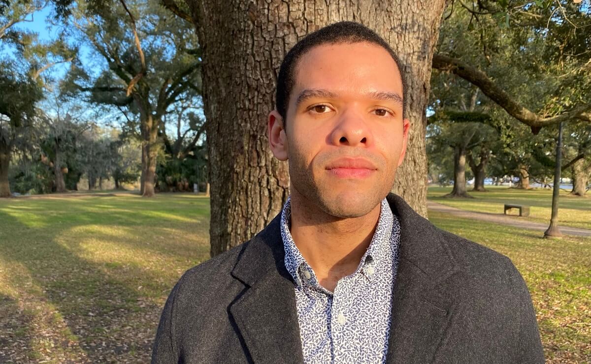 Photo of Miles Labat outdoors in what appears to be a large park, with recently mown grass and a concrete path on the far right that leads to a copse of trees in the background. Miles is wearing a button down shirt with a fine pattern of navy blue vines against white, and a dark gray wool overcoat. He is standing directly in front of a tree, and the warm glow of the sun on his face plus the long shadow of the tree behind him suggest late afternoon on a clear day.