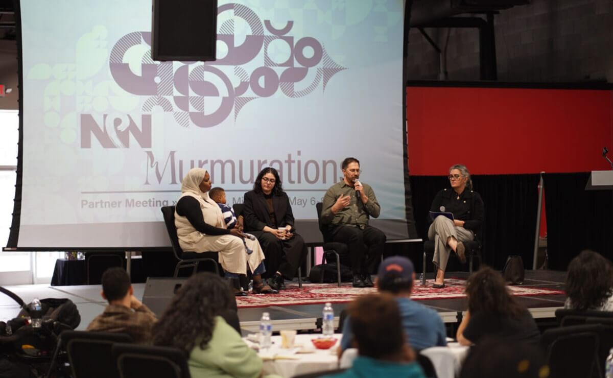 Detroit-area artists Layan Srour, Raphique Barakat, and Khadega Mohammed are seated onstage in front of an audience for a discussion moderated by AANM’s Director Dr. Diana Abouali. Behind the artists there is a projector screen with NPN’s logo, a graphic image, and information about the Partner Meeting, including the name of this year’s theme, “Murmurations.” Photo by Paisley Sutton, Winnie Media.