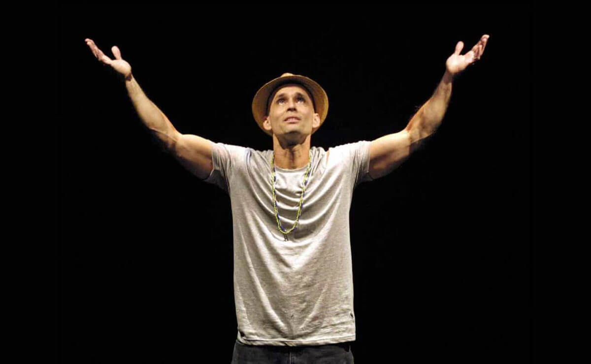 A light-brown-skinned man wearing a hat, a long beaded necklace, and a gray short-sleeved t-shirt stands against a black background and looks up as he raises his outstretched hands with the palms facing up.