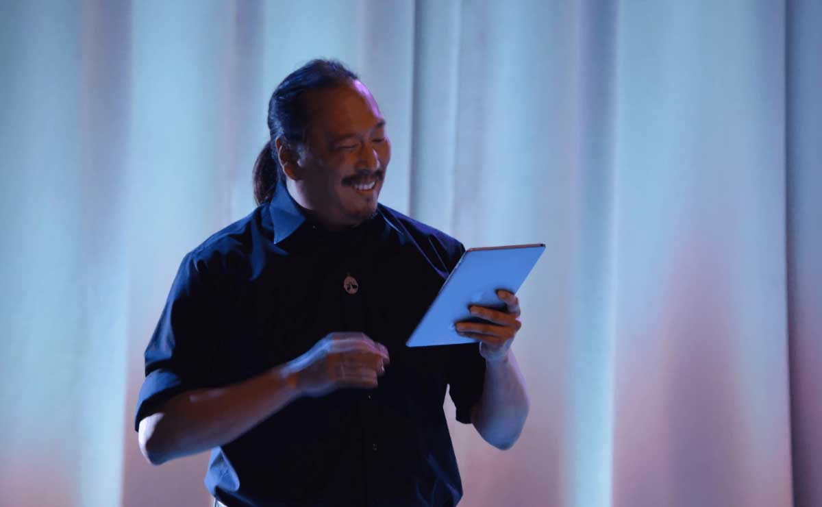 A photograph of Mendel Lee standing in front of a white curtain and smiling as he looks down at an iPad that he holds in his left hand.