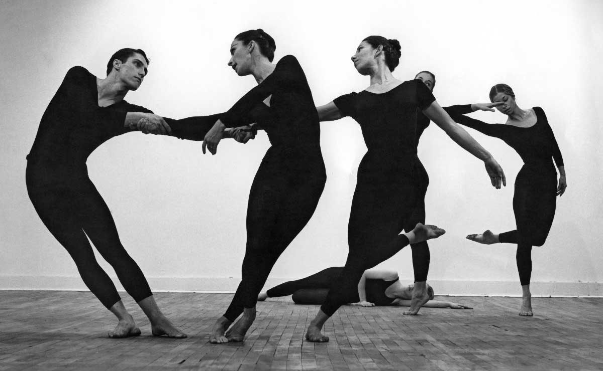 Black and white photograph of six dancers of various genders and skin tones and wearing black body suits performing on a bare wooden floor against an empty white wall. 