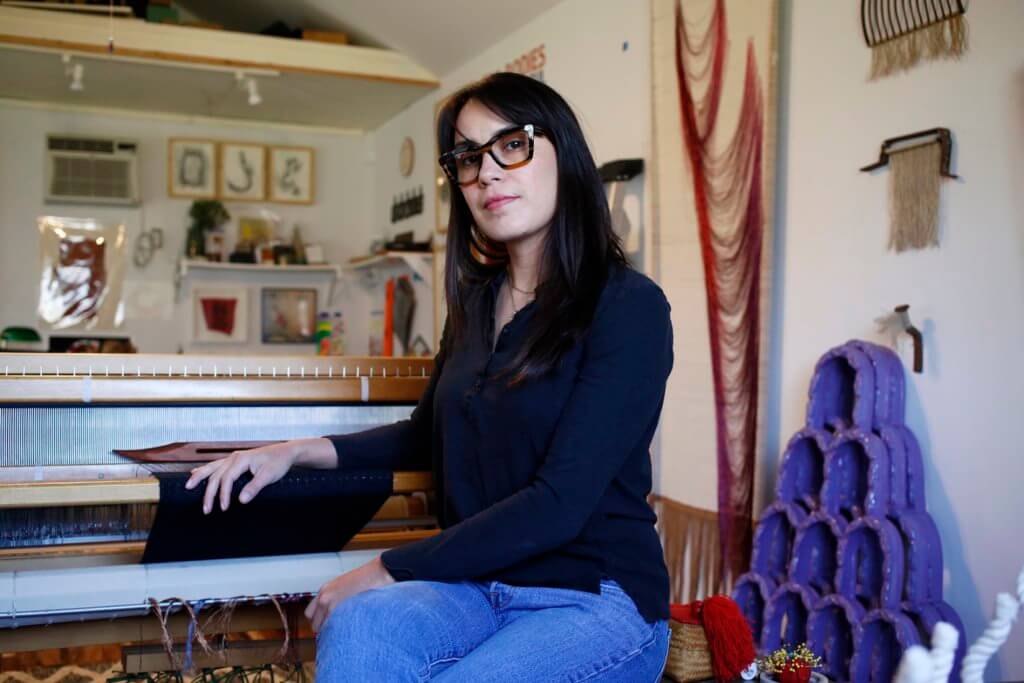 A medium-skinned woman with long dark hair, bangs, and thick-framed glasses sits with her right arm resting on her 4-shaft jack loom. She wears a black, long-sleeved shirt and jeans with her legs crossed at the knee. She is seen among the many items in her studio which include a small red pincushion, a woven box, and various artworks hanging on the walls behind her.