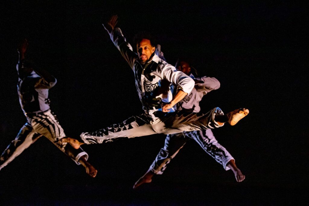 Three Black men leaping with arms and legs stretched out to the side. They are wearing white shirts and pants that have been painted/decorated with black ink.