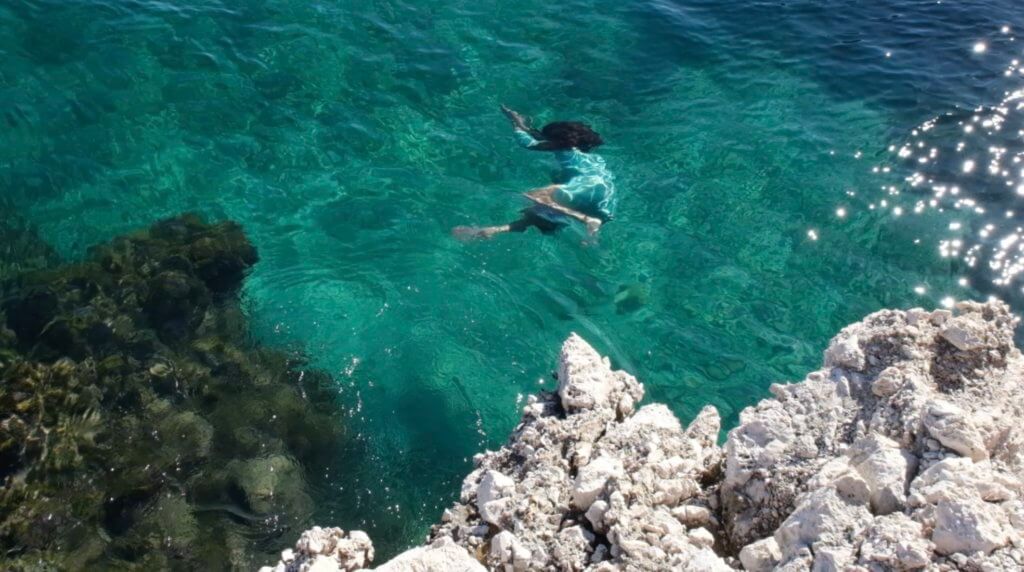 A Palestinian woman can be seen below the surface of turquoise water. Her spine is curved forward, knee in, arms above her brown hair. She is wearing a turquoise thobe that reflects sunlight. There is a jagged white stone on the bottom right of the image, and on the bottom left a stone underwater covered in green plant life. Sunlight is sparkling on the water.