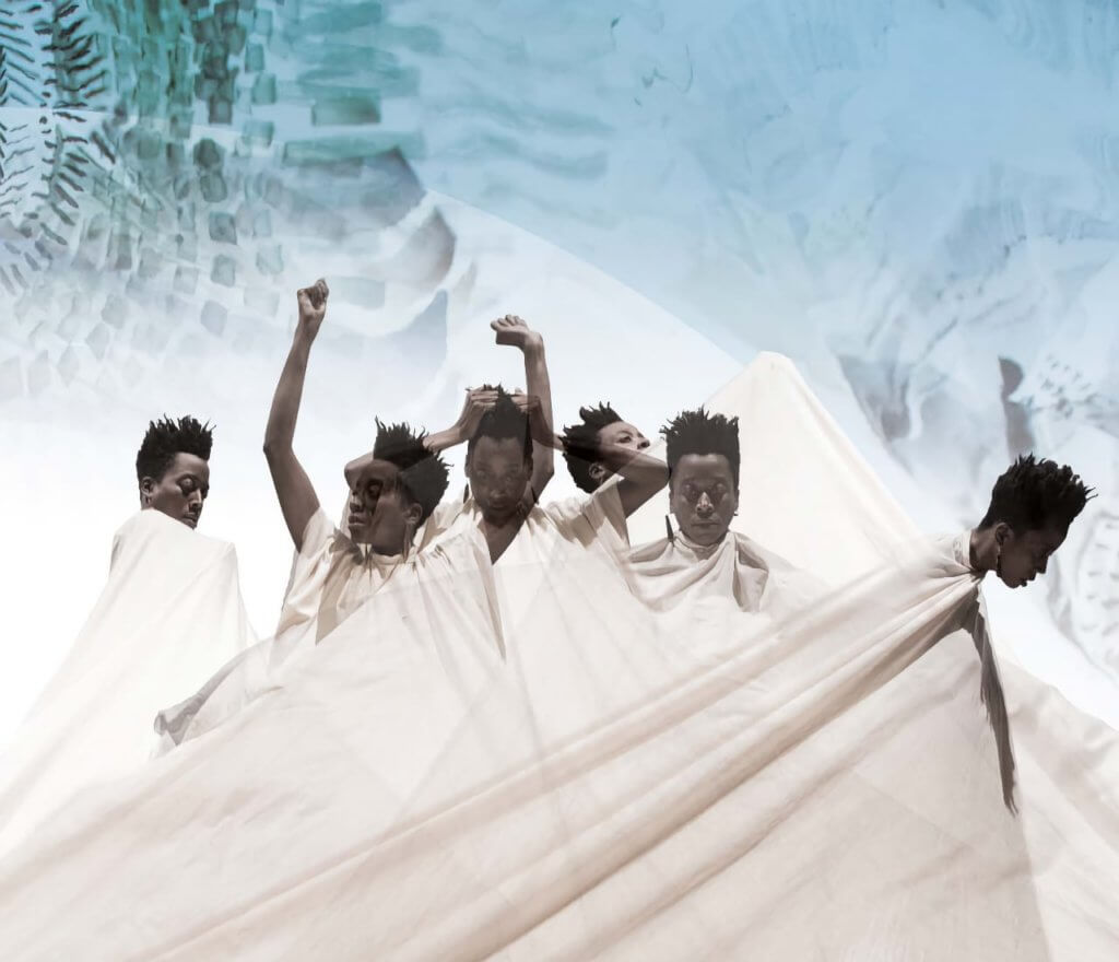 An African American woman with a short, tapered haircut is draped in a long white barbering cape. She is in front of a white, light blue, gray, and indigo background. Six moments of her dancing with the cape are overlaid to form a composite from left to right. She performs the following: she is wrapped in the cape, extends arms up with hands in loose fists and looks to the right, looks down and pulls her hair, pushes the cape up from underneath, calmly looks down and forward, then pulls away and to the side.