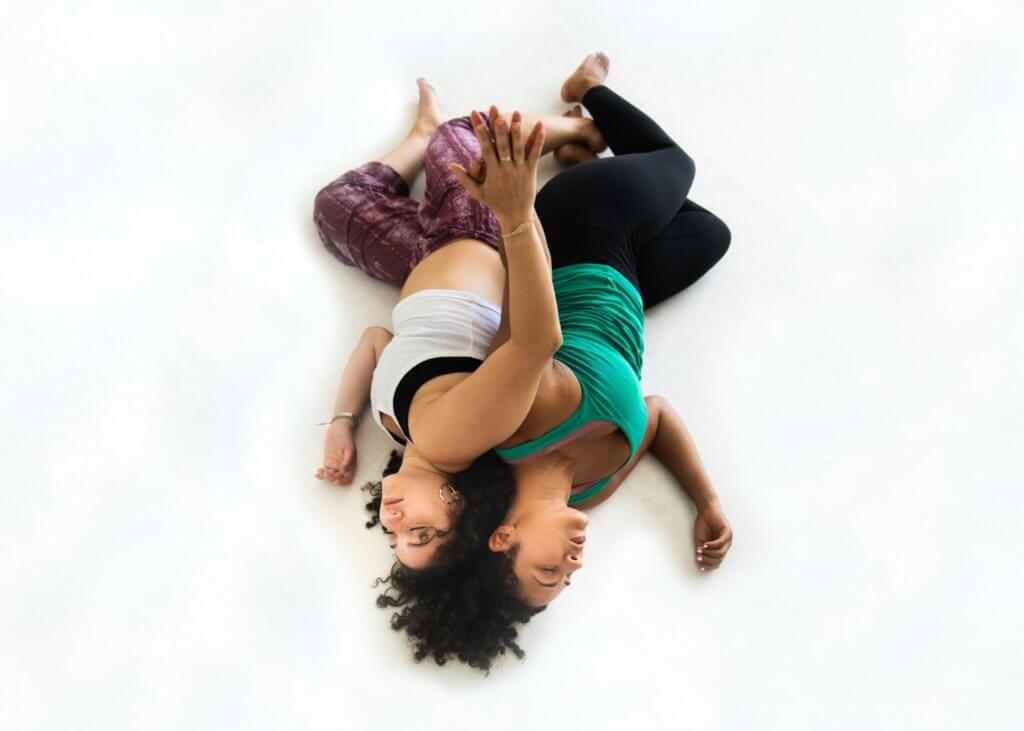 A medium-light skinned Latina woman, wearing a white tank top and purple pants, is on the floor back-to-back with a medium skinned Black woman, wearing a green tank top and black pants. Their eyes are closed, heads touching with brown curly hair spreading over both the tops of their heads (making it unclear whose hair it is). Their arms are entwined, palms joined reaching upwards.