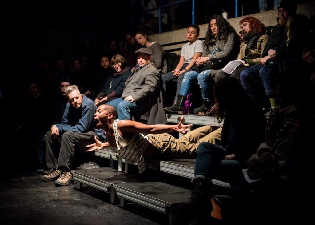 A brown-skinned non-binary person lays on a theater seating riser, surrounded by a sitting audience, and lifts their torso with arms outstretched and mouth wide.