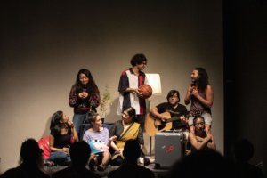 8 artists of Pan-Asian descent are gathered on a small platform in front of the audience. The performers are sitting and standing. They wear casual clothes and are talking to one another. One person plays a guitar, and one person is huddled in a corner by a suitcase.