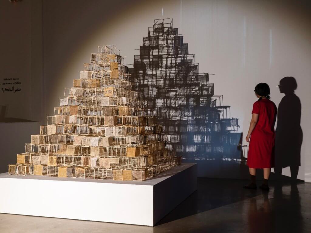 A brown woman in her late forties, artist Rehab El Sadek, wearing glasses and red dress. Looking peacefully at a huge sculpture comprised of hundreds of individual wooden cubes modeled after one of the pyramids of Giza. The structure is dramatically lit, creating a shadow that extends the volume and aspect of the work.