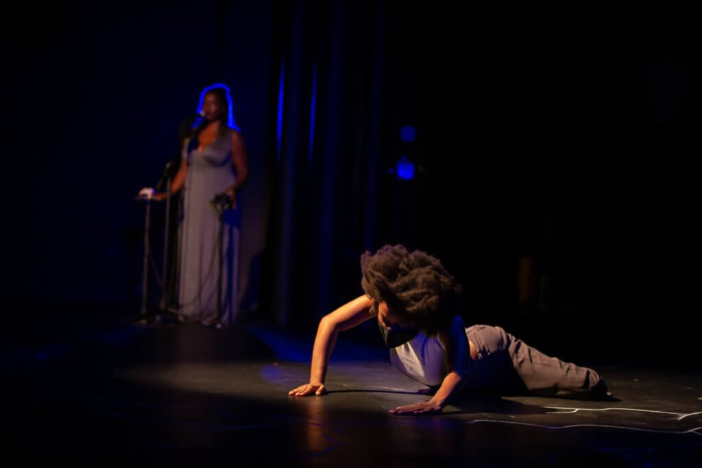 A dark-skinned woman is pushing her body up from the ground. Her arms are separated from the floor, and her head also erect looks to the floor although the lower half of her body remains connected to the earth. In the far back corner, a dark-skinned woman in a long gray dress stands behind a microphone, illuminated by a blue light.