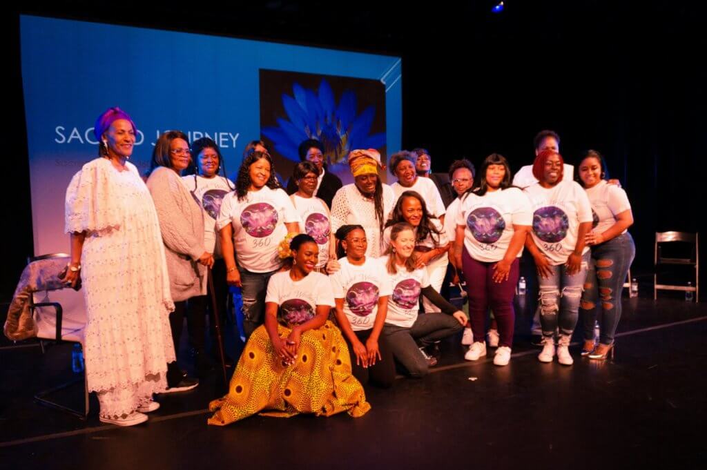 A group of 20 self-identified woman, majority Black, wearing white shirts with Sacred Wellness printed on them, standing onstage in front of a backdrop of a screen with the words “Sacred Journey” and a lotus. They are close together and smiling into the light, glowing from their performance.