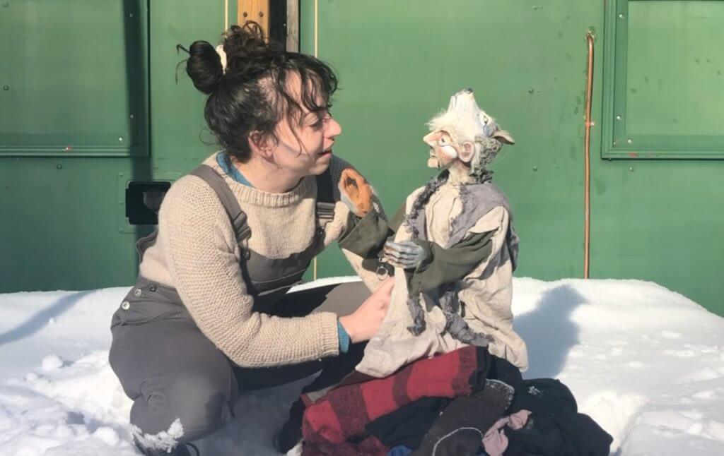 A light skinned woman with dark hair looks at a puppet. The puppet is an old woman with the face of a wolf on top of her head perched on the top of a pile of laundry. The two characters sit in the snow in front of a green background. Both are wearing a combination of grey and beige clothing. They are lit by the bright sun of a winter’s day.
