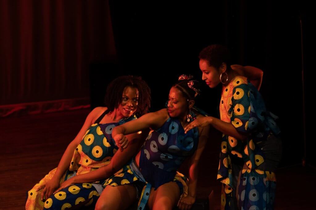 3 medium-brown skinned black women are pictured in sisterly love leaning into one another in dance gestures. Each woman wears an individual rendition of a of a yellow, orange, and blue African print fabric costume showing a repeating pattern of black notch edged spheres surrounded by concentric circles and dots. The woman on the left is seated, smiling, and leaning onto the seated woman in the center. She (center) is looking at her wrist in a gesture of checking time on a watch as she leans onto the woman on the right who is slightly higher looking on with interest.