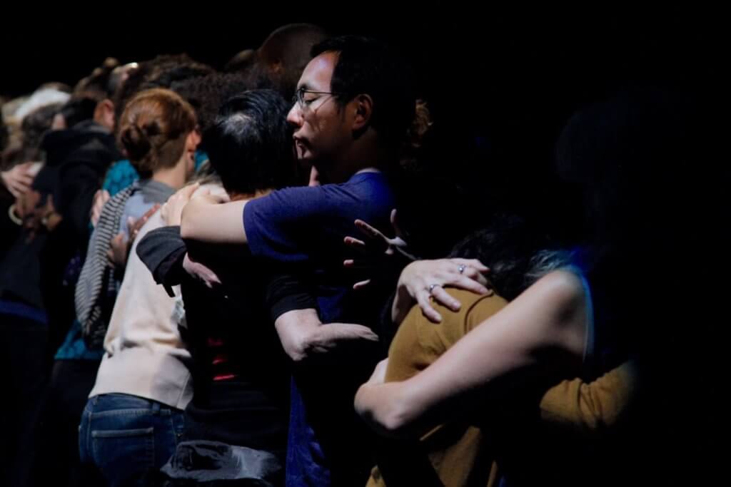 People hugging on a partially illuminated stage. Closer to the camera, only the arms of two people embracing are visible, next to them two other humans hug, and next to them another couple, creating a line of paired embraces. The person hugging in the center is most clearly discernible and is medium-skinned with short cropped black hair and wearing an indigo blue t-shirt. The end of the line is not visible.