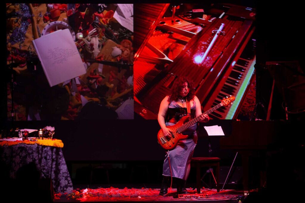 Brown woman with curly black hair stands on a stage covered with a Persian red rug in a blue dress, black and red corset, and black boots. She is holding a bass guitar and singing. To the right is a red baby grand piano. To the left is a table draped in black lace floral cloth. Behind her is a diptych video of the inside of the piano and tabletop. A sword lies on the piano. A notebook with a poem for her mother is open on the tabletop, surrounded by petals, skulls, and other femme treasures, covered with yellow shawls.