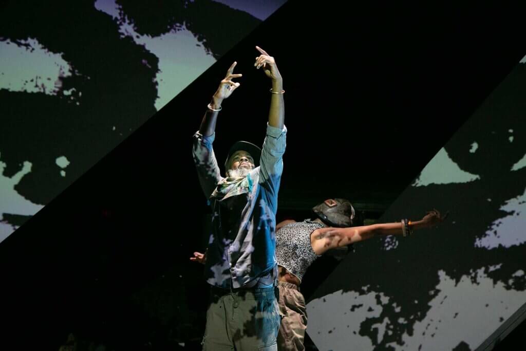 A Black man stands, looking up, arms stretched upwards, hands partially open, extending from his wrists. His pointer fingers are the furthest extended. He wears a cap; bracelets; mixed blue/gray/yellow scarf; gray/blue quarter-sleeve button shirt; and gray pants. Peeking out from behind him, a Black woman is dancing, and leaning her head and torso backwards. Both of her arms are outstretched, one overhead elongating the lean of her torso. She wears an African wooden mask, black/white tank top, bracelets, and brown pants. They are framed diagonally by a hyper-close projected image of a gray/green tone face, just the eyes identifiable.