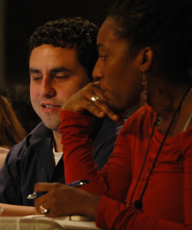 A close-up shot of two people at a meeting. In the foreground, a woman with dark hair, wearing a red long-sleeved shirt, has her hand to her chin in a thoughtful gesture as she holds a pen. Behind her, a clean-shaven Alec De León looks down toward the table, wearing a dark blue shirt over a white undershirt. They appear to be engaged in conversation.