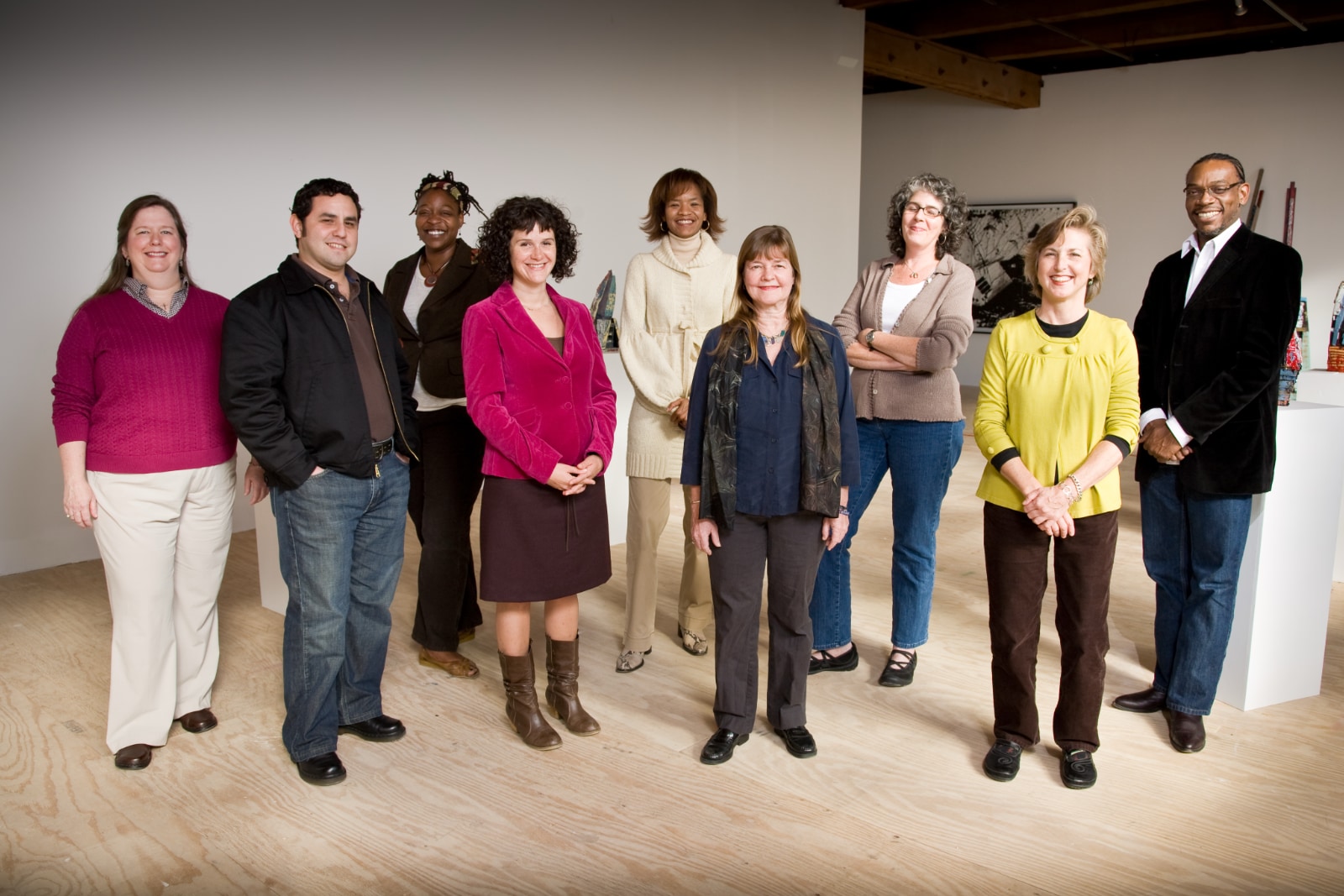 A group of nine people pose for a formal staff portrait in a brightly lit, open-plan art gallery or office space with light wooden flooring and white walls. From left to right: a woman in a magenta sweater and tan pants; Alec De León in a black jacket and jeans; a woman with dark hair in a dark blazer; a woman in a vibrant pink velvet blazer and dark skirt; a woman in a cream-colored turtleneck sweater; a woman in a dark shirt and patterned scarf; a woman with curly hair wearing a brown cardigan and jeans; a woman in a bright yellow top and brown pants; and a man in a black velvet blazer and jeans.