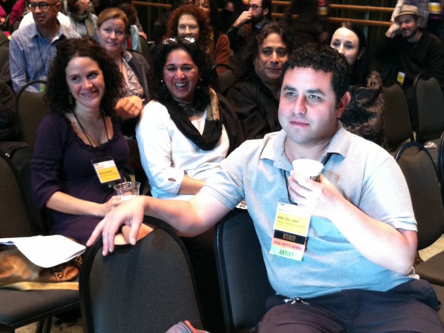 A medium shot shows a diverse audience seated in a conference room. In the foreground, Alec De León, wearing a gray polo shirt and holding a cup, looks forward. A long name badge hanging from his neck includes the words "Staff" and "Artist." Two women are seated nearby in the row behind him. One wears a dark purple dress, and the other, wearing a white top and black scarf, is laughing. The background is filled with other attendees sitting in rows of chairs.