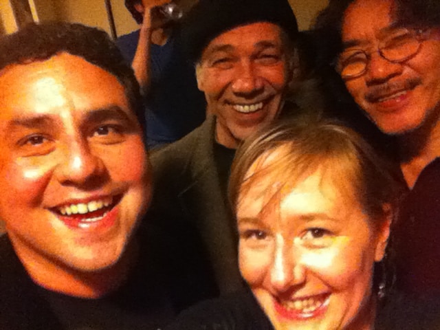 A close-up selfie shows four people smiling together indoors. In the foreground on the left is Alec De León, a young man with dark hair, and in the foreground on the lower right is a woman with light brown hair. Behind them stand two men; one in the center wears a dark beret, and the other, on the right, has a slight mustache and wears glasses.