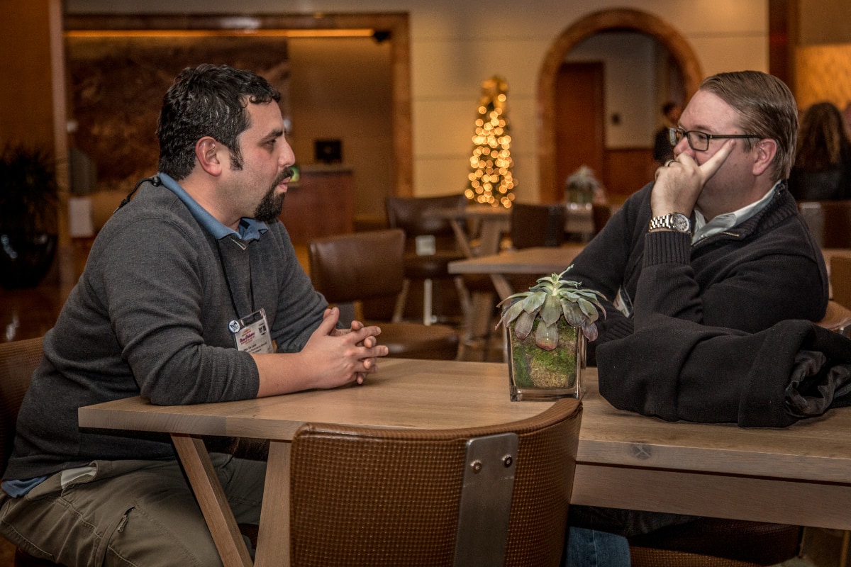 Two men sit across from each other at a wooden table in what appears to be a hotel lobby or event space. The man on the left, Alec De León, wears a dark gray sweater over a blue collared shirt and has a conference badge clipped to his clothing. The man on the right wears glasses and a dark sweater, resting his chin on his hand. A small potted succulent sits on the table between them. In the blurred background, there is an arched doorway and a lit Christmas tree.