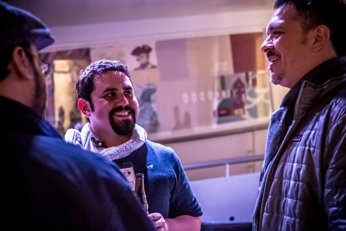Three men are engaged in conversation indoors. Just left of center of the image is Alec De León, with a dark goatee, a dark shirt, and a light-colored scarf. Alec is smiling at the man on the right, who is taller than the other two men in the photo. He has a much fainter goatee and is wearing a gray jacket, and he is shown in profile. A third man, seen from behind and wearing a cap, is partially visible on the left. The scene is illuminated with purple-toned lighting.