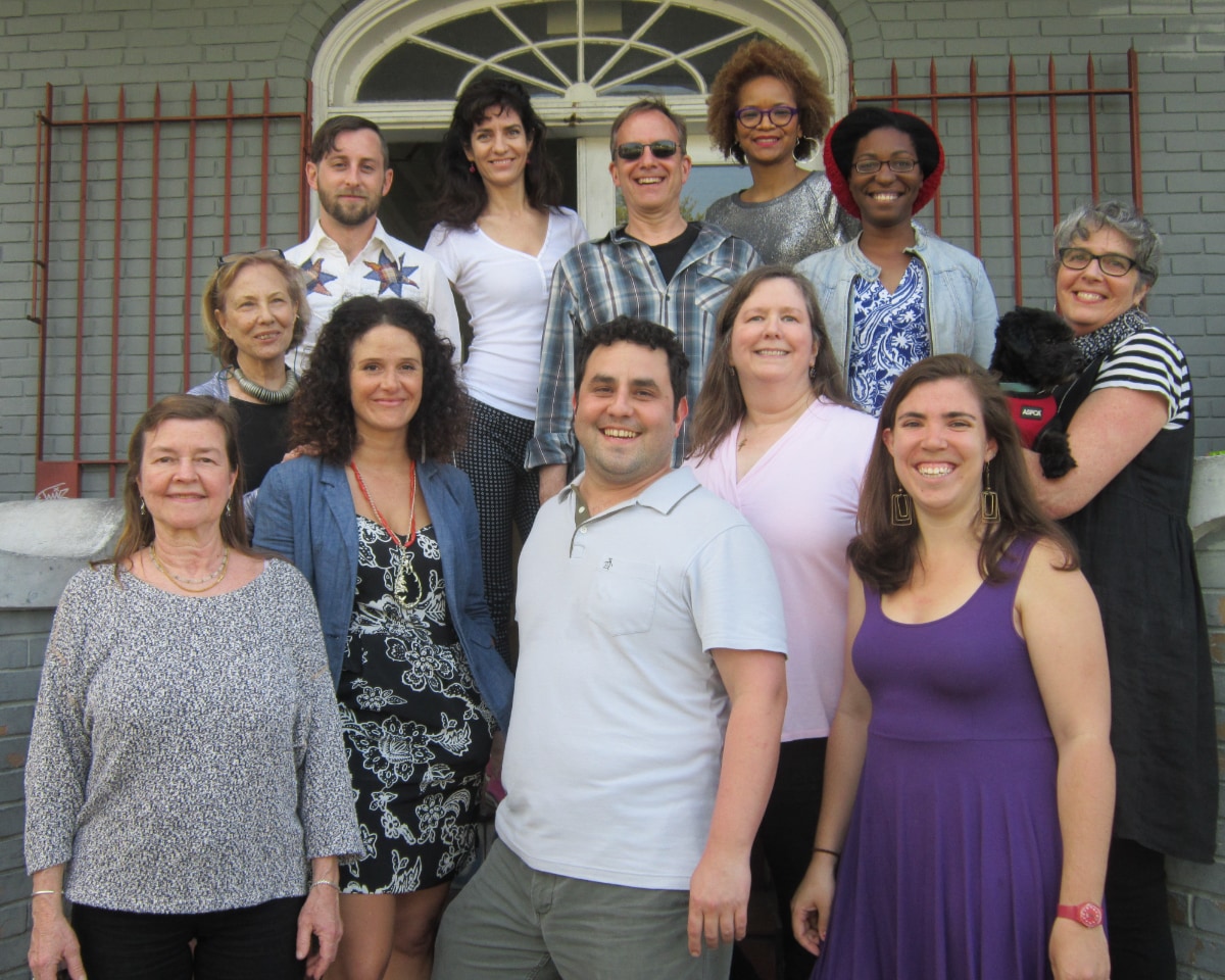 A group of twelve people pose for a formal portrait on the steps of a brick building. In the front row, from left to right: a woman in a gray patterned top; a woman in a blue blazer over a floral dress; a clean-shaven Alec De León in a light gray polo shirt; a woman in a pink top; a woman in a purple dress; and a woman in a black-and-white striped shirt holding a small black dog in a red harness. In the back row, from left to right: a woman in a black top with a chunky green necklace; a man in a white star-patterned shirt; a woman in a white v-neck; a man in a plaid button-down; a woman with curly hair wearing glasses; a woman with a dark red beanie.