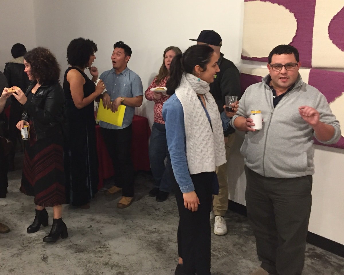 A group of people gather in an indoor space with white walls and a large, abstract purple-and-cream art installation. In the foreground on the right side of the image, near the wall with the abstract art, is a clean-shaven Alec De León wearing glasses, a gray fleece jacket with a zipper, and dark pants. Standing next to Alec and engaged in conversation with him is a woman with a black ponytail, wearing a blue denim jacket, a thick white scarf, and dark pants. Behind them, half a dozen other people are talking in small groups while holding drinks or small plates of food.