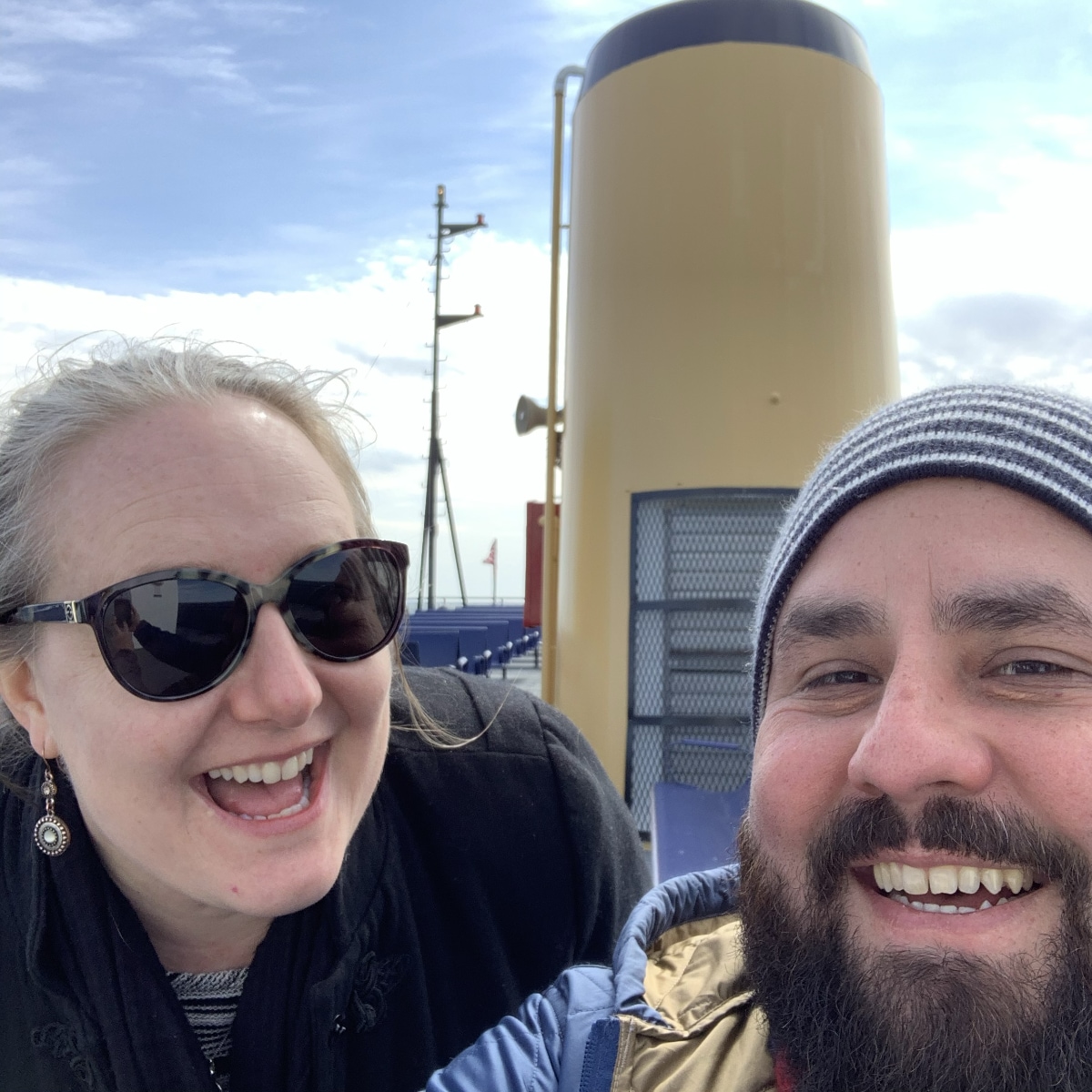 A selfie of a smiling woman with blonde hair wearing sunglasses and Alex De León, a man with a dark beard and a striped beanie. They are outdoors on what appears to be the deck of a ferry, with a large yellow smokestack visible behind them against a bright, partly cloudy sky.