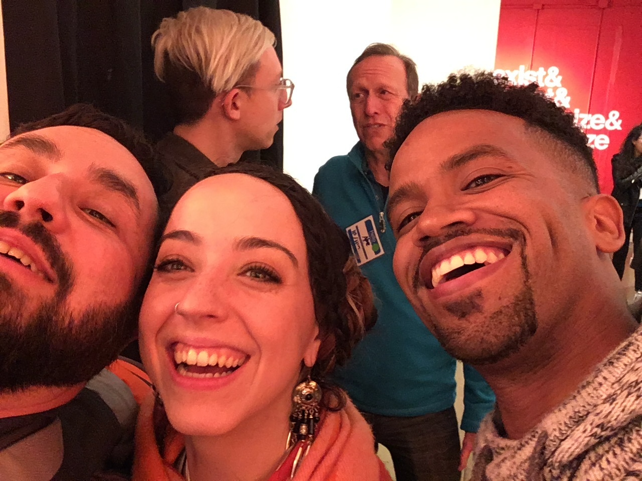 A group of people pose for a selfie in an indoor setting during an event. On the left, with his face partially cut off by the edge of the photo, is Alec De León, a man with a dark beard. In the center is a smiling woman with dangling earrings, and on the right is a man with a goatee. In the background on the far right, a partial red sign with white text is visible.