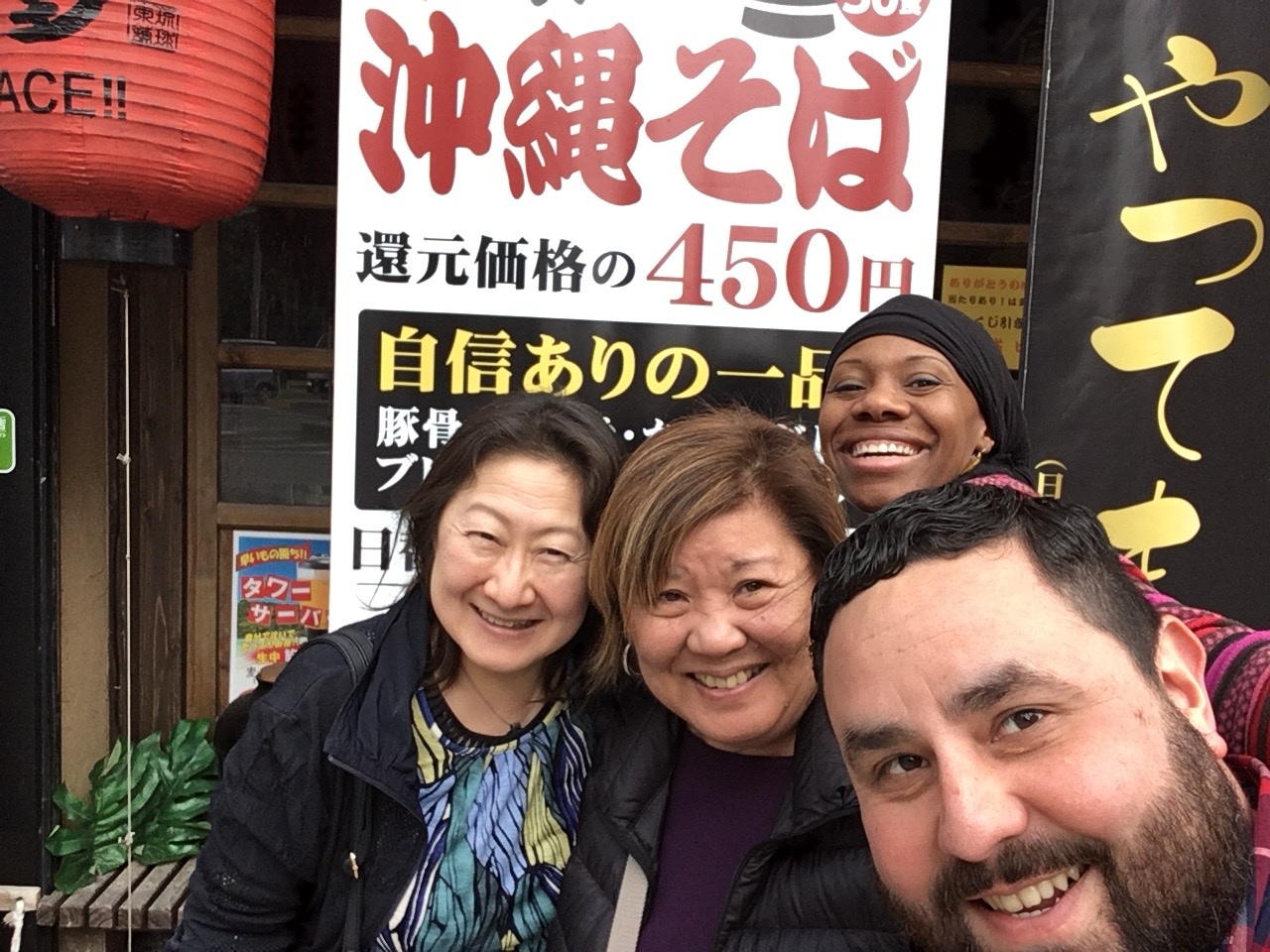 A group of four people take a selfie together in front of a shop sign with Japanese text. In the foreground on the bottom right is Alec De León, a man with a dark beard. Behind him and to the left are three women smiling for the camera: one in the back right wearing a black head covering, and two in the front left wearing dark jackets. A large red paper lantern hangs on the left side of the frame.