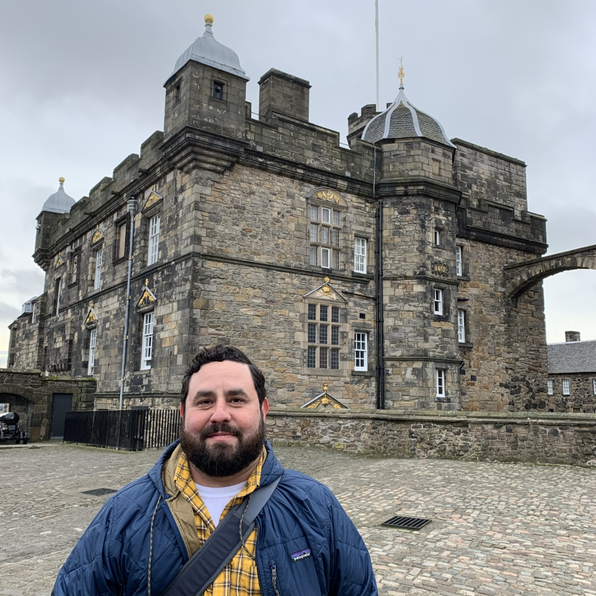 Alec De León, a man with a dark thick beard and wearing a yellow plaid shirt under a blue quilted jacket, stands in a stone courtyard. Behind him is a historic stone building within Edinburgh Castle, which features arched windows and decorative gold elements above the windows.