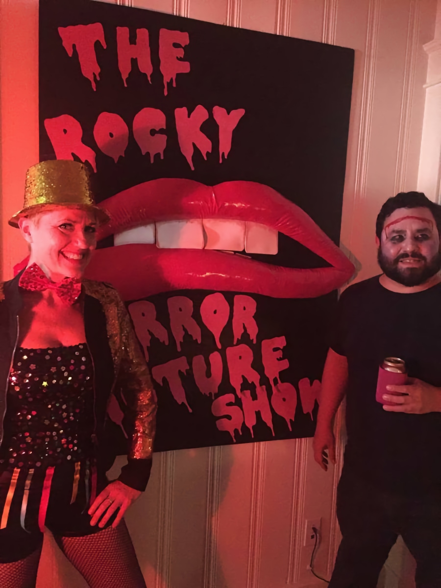 Caitlin Strokosch and Alec De León, in costume as characters from "The Rocky Horror Picture Show," pose for a photo in front of a large poster for the movie featuring the iconic red lips from the opening credits. On the left, Caitlin is dressed as the character Columbia in a sparkly gold top hat, jacket, and fishnet stockings. On the right, Alec is dressed as the character Eddie, with white face paint, dark shadows around his eyes, and a long horizontal incision mark across his forehead.