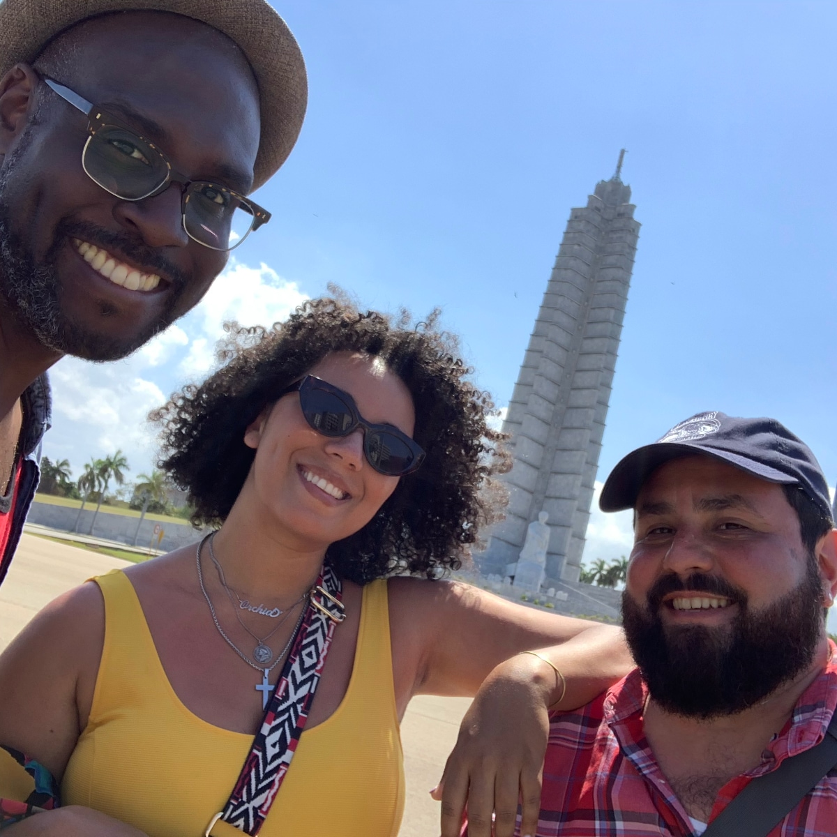 A selfie of three people smiling outdoors with a tall, slender stone monument in the background against a clear blue sky. On the left, a man wears a hat and glasses. In the center, a woman with curly hair wears a yellow tank top and sunglasses. On the right is Alec De León, a man with a dark beard and wearing a baseball cap and a red plaid shirt.