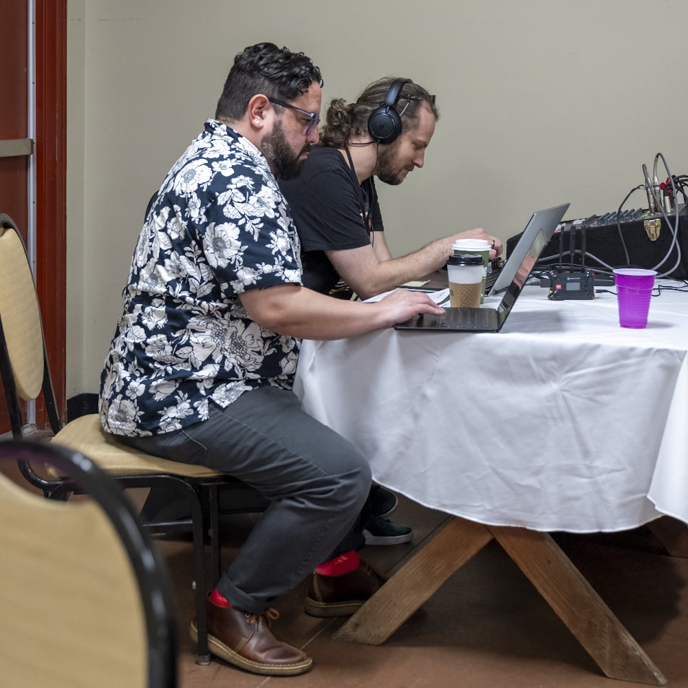 A bearded Alec De León and another man are shown in profile seated side-by-side at a table covered with a white tablecloth, working on laptops. Alec, in the foreground, is wearing a floral-patterned short-sleeve shirt and dark pants, and typing on a laptop. Beside him, a man with long hair and wearing headphones is also focused on a laptop, with a sound mixing console positioned further along the table.