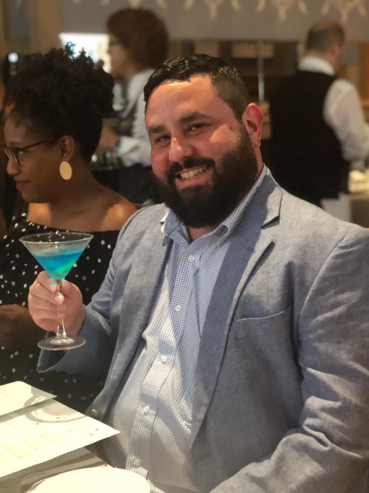 Alec De León, a bearded man in a light blue blazer and a checkered shirt, smiles while holding up a bright blue cocktail in a martini glass at a table. Beside him, a woman with short dark hair, wearing glasses and a black polka-dot dress, is partially visible. The background is a soft-focus restaurant scene with other patrons and staff.