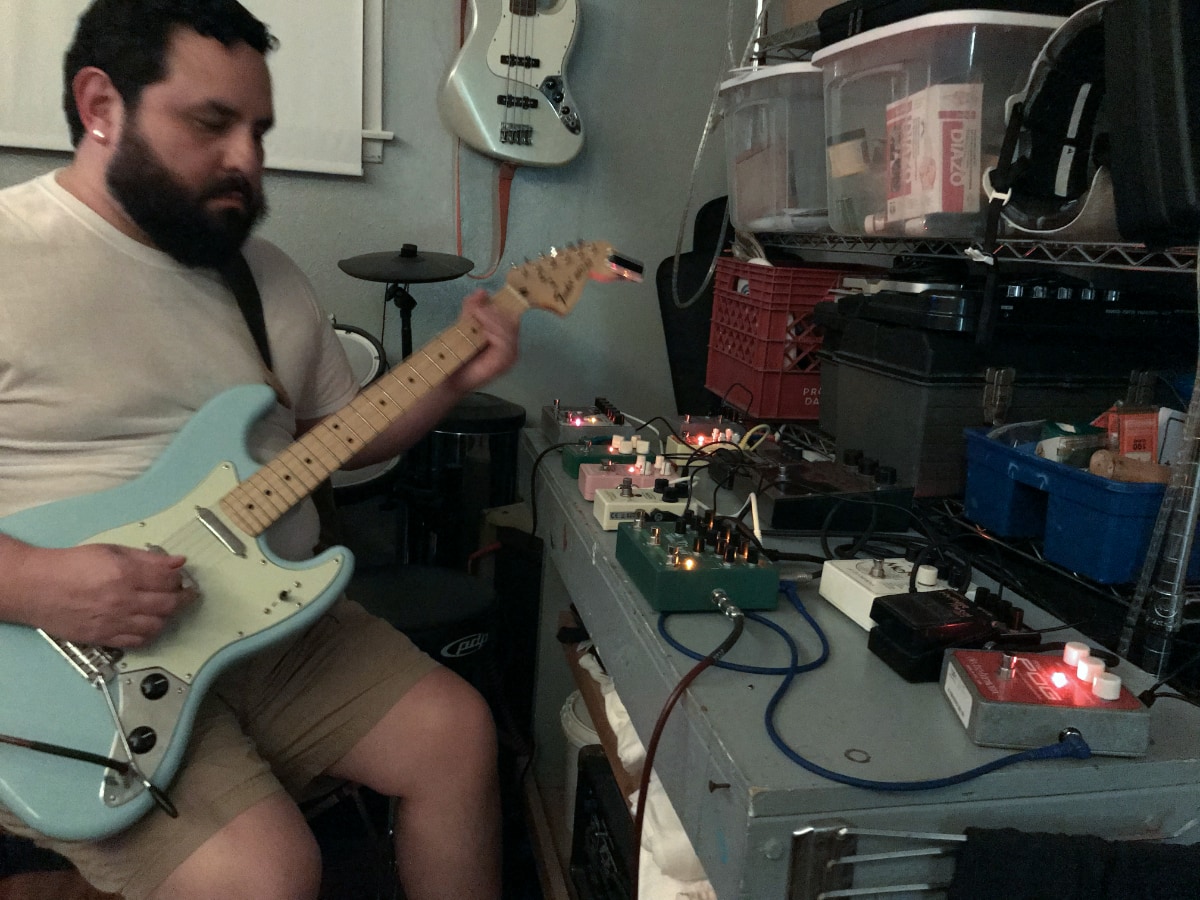 Alec De León, with a thick dark beard and wearing a white t-shirt and tan shorts, plays a light blue electric guitar while seated. On the workbench in front of him is a row of guitar effects pedals with glowing lights and connected by cables. A bass guitar hangs on the wall behind him, and various storage bins and equipment are visible on shelves to his right.