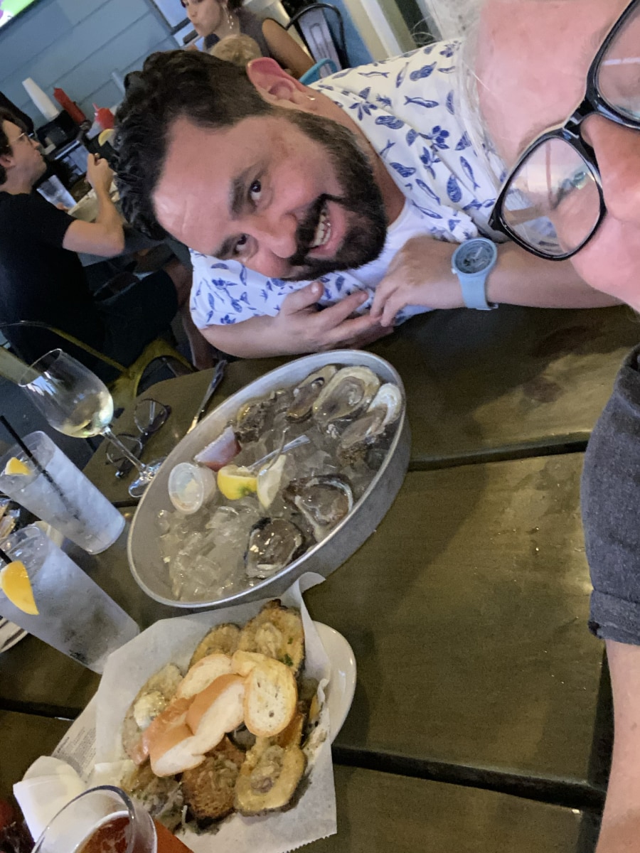 A close-up, top-down selfie shows a bearded Alec De León smiling at the camera while leaning over a table at a restaurant. A large metal platter filled with fresh oysters on ice is centered on the table. Next to the platter is a dish of cooked oysters topped with toasted bread slices. The photographer is partially visible on the right, wearing glasses.