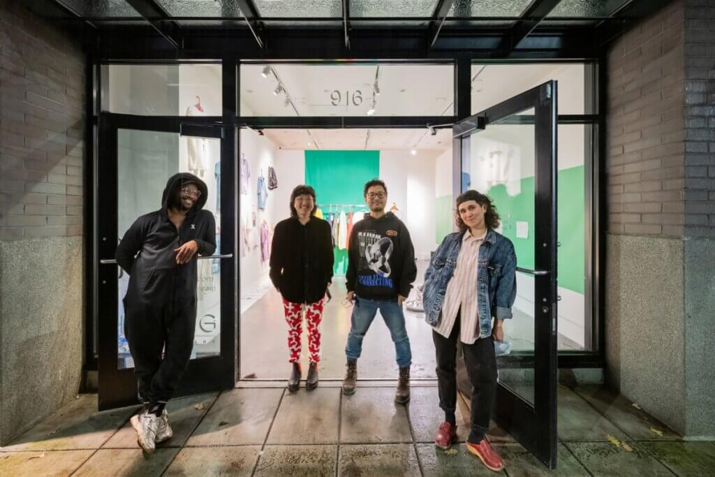 A group of four people stand in the open glass doorway and windows of a gallery. From the left, a Black person wearing a black jumpsuit, an Asian person wearing a black blouse and red and white pants, an Asian man wearing a graphic sweatshirt and jeans, and a white woman wearing a denim jacket, striped blouse and black jeans are looking at the camera, smiling. Behind them you can see green screens lining the walls, clothing on racks and on the wall in the brightly lit gallery space.