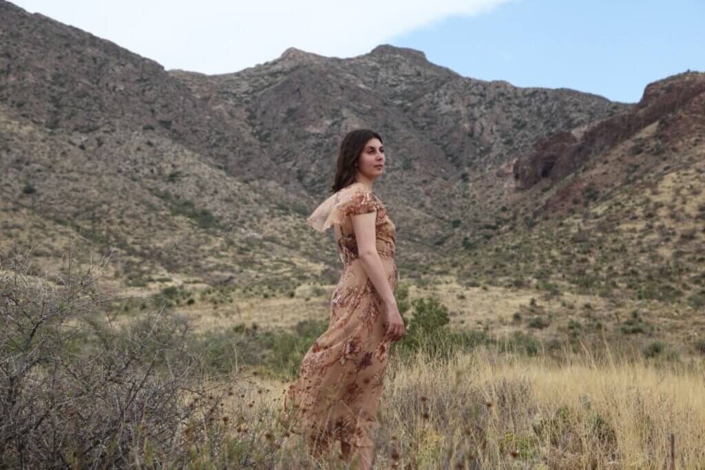 A woman in a brown floral dress facing sideways, looking over her right shoulder, in front of towering desert mountains. The wind is blowing her dress slightly, blending it into the desert shrubs at her feet.