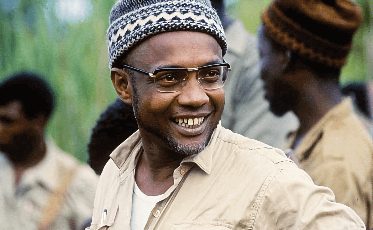 Portrait of Amílcar Cabral smiling. He is wearing glasses and a patterned knit cap, and is dressed in a light-colored shirt with the collar slightly open to reveal a white undershirt. The background is softly blurred, showing other individuals and greenery, suggesting an outdoor setting. The man’s expression conveys warmth and approachability.