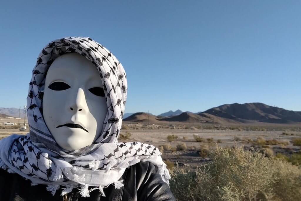 A masked figure with a death-white face, solid black eyes, and a down-turned mouth, wearing a black leather jacket and a keffiyeh (black and white patterned headscarf). The figure stands in front of a desert landscape with blue sky, patchy desert greenery, and distant mountains.