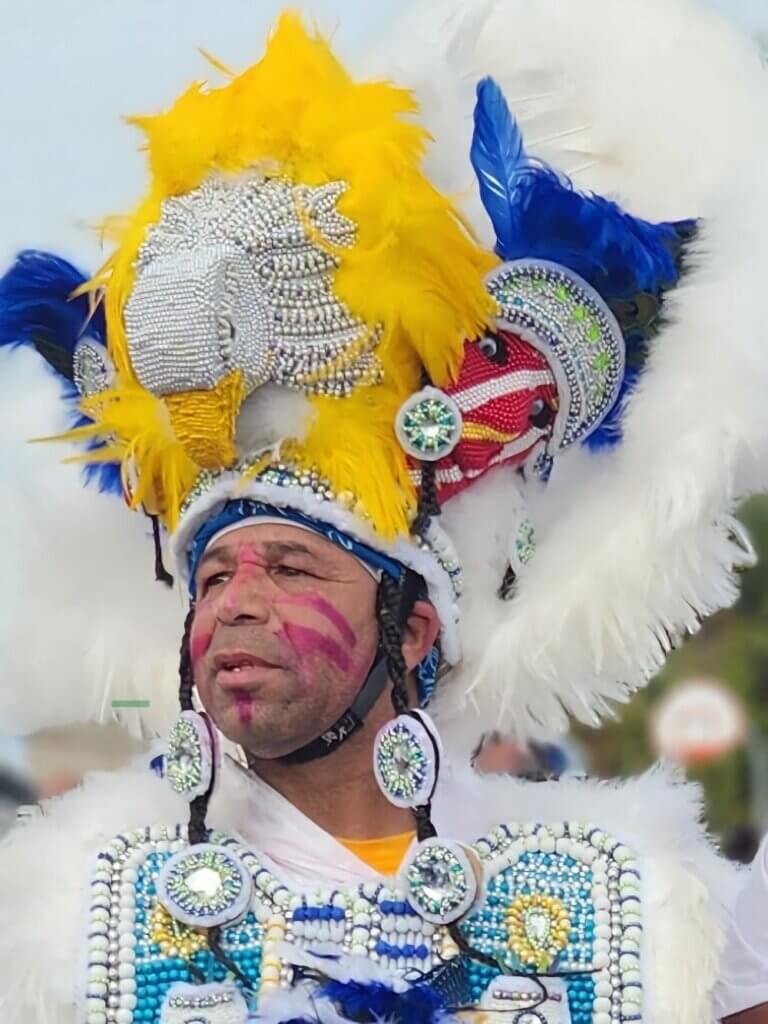 An image of a medium-skinned man looking off into the distance wearing a large white, yellow, and blue feathered headpiece with the head of a white eagle atop it. He has red paint on his face and wears a white and blue beaded and feathered Black Indian suit.