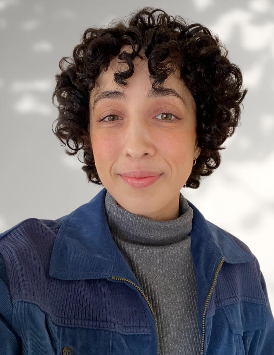 The photo depicts Art2Action's General Manager, Gabi Figueira, who has medium light skin and short, dark curly hair. She is wearing a grey turtleneck and a navy blue jacket against a white, sun-dappled background.