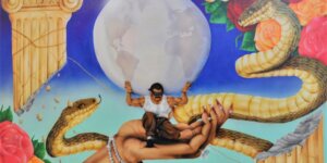 A symbolic and surreal illustration of the myth of Atlas. Against a blue background, a muscular Latino man with dark hair and a mustache crouches in the palm of a woman's bejeweled hand. He is wearing a white muscle t-shirt and brown pants, and he is blindfolded with a blue bandana. Upon his broad shoulders is a white and gray representation of Earth approximately three times larger than the man, with North and South America visible on the left half and Europe and Africa visible on the right half. The man's hands are bound with gold cords that extend to the top half of a broken pale yellow Greek column in the upper left corner of the painting, and to the bottom half of a broken pale yellow Greek column in the bottom right corner of the painting. Pink, red, and orange roses in full bloom fill the adjacent corners. Three giant, brownish-green snakes, with bellies the color of the Greek columns and heads as big as the man's chest, fill the space between the Earth and the four corners of the painting. One of the snakes is coiled around the wrist supporting the outstretched woman's hand; another snake is rising up from the bottom of the painting directly beneath the hand; and the third extends from behind the lower right column to behind the man, then up and over the roses in the upper right corner.