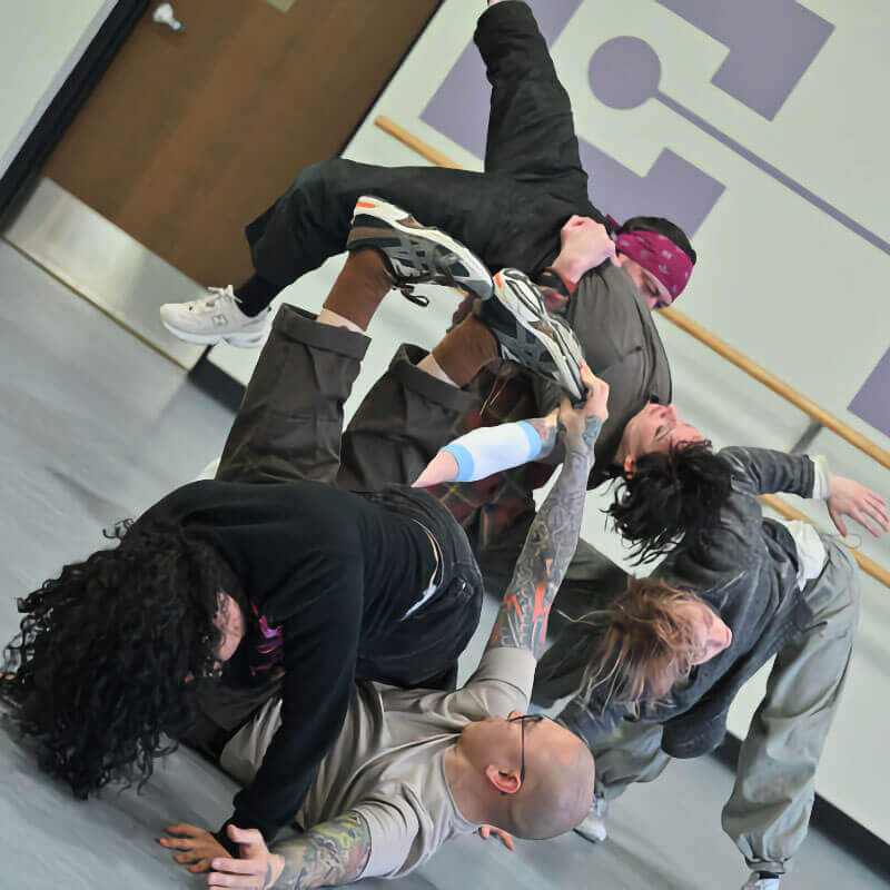 In a dance rehearsal space, five dancers of various ethnicities and genders, all wearing loose fitting gray or black clothing, pose together with limbs intertwined and bodies bent in various unusual ways. The photo is tilted, adding to the visual chaos.