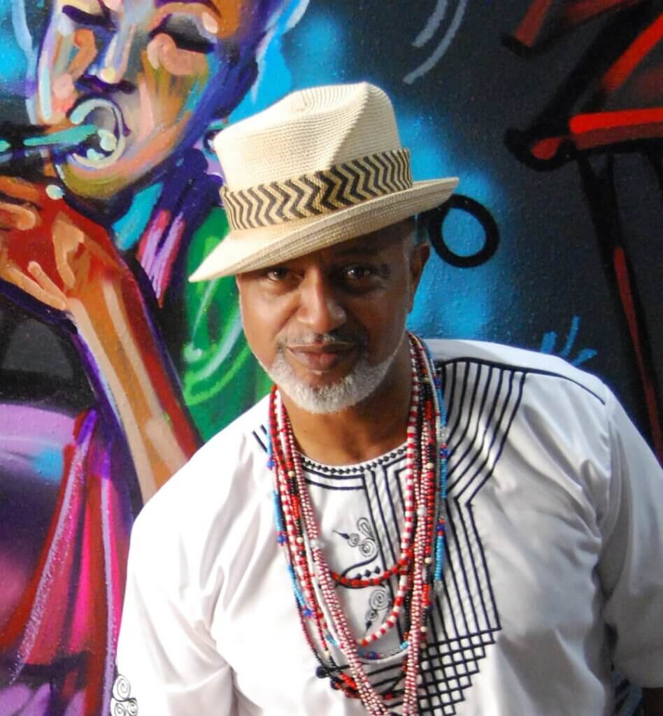 The image is of a medium, dark-skinned African-American man with a light gray mustache and beard. He is wearing a beige straw fedora, titled to the side, and a white kaftan with navy blue embroidery around the collar. Behind him is a colorful mural featuring a musician playing a horned instrument.