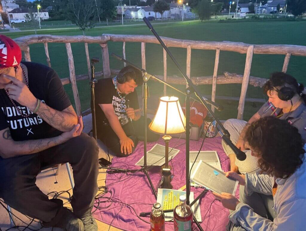 Four actors perform a dramatic reading of the original script of La Fête Du Petit Blé Ou L'héroïsme De Poucha-Houmma at dawn at Nanih Bvlbancha. Three of the actors are sitting on blankets, and the fourth is sitting in a folding chair. There is a lamp between them and they are looking down at their scripts. Behind them is an irregular fence made out of rough-hewn branches and thin logs.